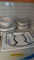 Various CorningWare bakeware pieces arranged on counter, including oval and square casseroles, some stacked, with glass lids and a metal lid holder