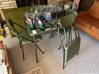 Overall view of green vinyl padded folding table with four matching folding chairs around it. Table surface has various glassware and kitchen items on top. Chairs and table legs are metal framed.