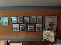 Wide shot showing 11 framed color Gary Patterson golf prints arranged on a cork wall and 1 print leaning on shelf plastic wrapped.