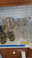 Multiple glassware items including frosted mugs, clear glass mugs, a decorative bowl with handle, clear wine glasses, and plastic holders in green color on a kitchen countertop.