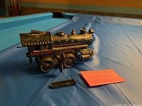 Photo of the black metal wind-up train locomotive from a side angle showing front wheels and detached rectangular piece on a blue surface.