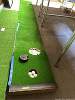 Putting green mat laid out on carpeted floor showing two golf cups and three balls in one hole. Blue tape and rolled end of green turf visible.