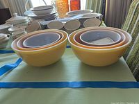 Two sets of three nested Pyrex bowls each, placed side-by-side on a table with other kitchenware in the background. Bowls shown in yellow, orange, and brown autumn colors.