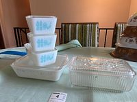 Stacked view of three small vintage milk glass Pyrex dishes with blue rooster and corn pattern and lids on top of the large rectangular milk glass dish with matching pattern.