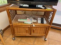 Front view of rattan cart with two cabinet doors, top shelf holding TV, and various cables and remotes on shelf.