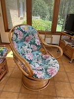 Front and angled views of a rattan swivel chair with floral cushions on a tiled floor in a sunlit room.
