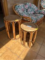 Two round rattan stands with woven tops placed side-by-side on tile floor beside a rattan floral cushioned loveseat.