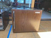 Mini fridge with wood grain finish on side and front, vertical silver handle, placed in bright tiled indoor area.