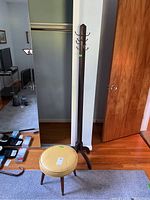 Full view of tall wood coat rack with stool in front, the coat rack has multiple hooks with one visibly broken, set on wooden floor indoors.