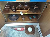 Wooden serving bowls, trays, and coasters arranged on wooden shelf and floor showing variety of shapes and sizes.