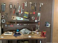 Wide view of pegboard with various hand tools hung, including wrenches, trowels, saw, tapes, stapler, and other small hand tools above a wooden workbench with other items present.