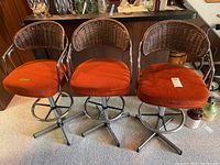 Front view of three matching vintage Cosco bar stools with metal bases, orange upholstered seats and rounded wicker backs showing wear.
