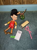 Full lot photo showing Pinocchio doll, wooden dog, Zell bank and key with pink tool on carpeted floor.