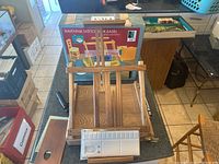 Wooden Ravenna Sketch Box Easel in folded position on counter, original box behind it. Attached white plastic palette, brass hardware visible.