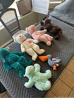 Six assorted TY plush toys laid out on a gray quilted surface near a window and chairs, showing variety in colors and animals including teddy bears and a bird. Each appears to have original TY tags attached.