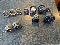 Image of a variety of 9 aviation gauges laid on a counter featuring round and double faced round gauges in black, with white lettering and needles.