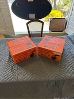 Two unopened orange Super Marel bocce ball boxes on a folded quilted gray cloth surface, showing front and side views of the boxes.