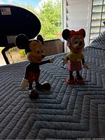 Two vintage Mickey Mouse and Minnie Mouse toys placed on a grey quilt with plantation shutters and outdoor background visible