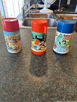 Three collectible thermoses by Aladdin Industries displayed on a counter, showing front graphics and different colored lids red, orange, and blue.