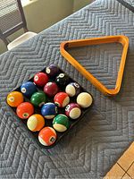 Top-down photo showing 15 billiard balls organized in a tray and adjacent orange triangular billiard rack on a gray quilted surface.