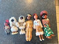 Six vintage dolls on dark surface showing diverse traditional and cultural outfits, with some dolls featuring eyes that open and close. Dolls show wear with one having a detached leather head.