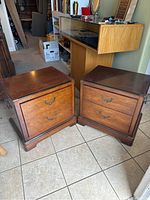 Pair of two wooden nightstands shown from front, displaying two drawers each with metal handles, placed on tile floor in a cluttered room.