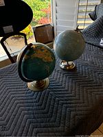 Two small globes placed on a quilted black surface near window light. One globe with teal ocean and brass-colored base, the other with pale blue ocean and light metal base.