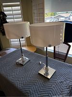 Two identical table lamps with white drum shades and chrome bases photographed side-by-side on a gray padded surface inside a room near windows