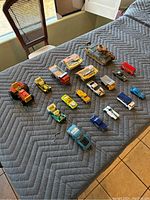 Top view of 18 toy vehicles laid out on grey padded blanket, showing variety of colors and types including cars, trucks, tank, and bus.