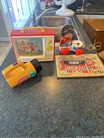 Lot of vintage toys on kitchen counter showing all items: TV music screen toy, wooden Drummer Boy pull toy, Playskool flashlight, and wooden farm puzzle.