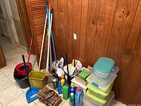 Wide view of cleaning lot showing spin mop, broom, dustpan, waste basket, iron, cleaning liquids, and other supplies arranged against a wood panel wall.