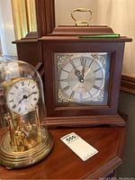 Both clocks on wooden surface; brass anniversary clock dome and wooden mantel clock side by side