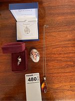 Full lot overview showing all four items on wooden surface: Swarovski Family necklace in box, amber pendant necklace, pin in burgundy box, and pill container with cherub decoration.