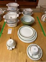 Overview photo showing 7 Corning Ware casserole dishes with lids, nested green Pyrex mixing bowls, and Corelle plates and cups on a wooden surface.