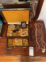 Overview of costume jewelry lot including opened wooden jewelry box with yellow velvet inside showing multiple costume jewelry pieces such as necklaces, bangles, pins, watch, earrings, and pendants alongside additional necklaces outside the box on a wooden surface.