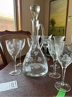 Overall view of etched glass decanter with stopper in the center surrounded by six matching etched wine glasses on a dining table.