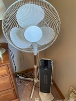 White floor fan and black DeLonghi ceramic tower heater side by side placed against a wall with cords visible