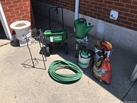 Photo of Scotts spreader, watering can, hose, metal plant stand, sprayer, and bag of manure on concrete patio with brick wall background. Concrete pot seen but not included.