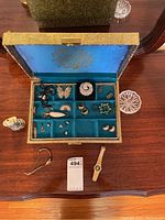 Open jewelry box showing assortment of earrings, pendants, pins, two watches, and other jewelry pieces arranged inside velvet-lined compartments