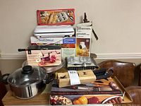 Kitchen lot with various items stacked including ravioli maker box, Seal-a-Meal vacuum sealer, apple peeler, cheese grater, pot with lid, BBQ tool set and bottle capper