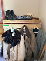 Three men's jackets hanging on hangers with a woven hat on top of cabinet above, boots and shoes visible on top surface