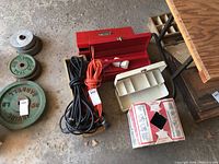 View of the lot arranged on floor showing two red metal toolboxes (one open), white metal tackle box, two coiled extension cords (one black, one orange), partial box of tying twine, and some small items.