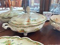Covered serving dish with floral patterns, lid, and three feet shown on wood surface with other dinnerware