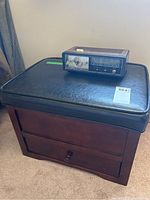 Photo of ottoman seat with black cushioned leather-like top and wooden base featuring storage drawer and a Realistic Chronomatic 9 clock radio placed on top.