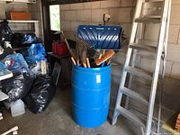 Overview of blue rain barrel filled with garden tools and 5 ft aluminum step ladder nearby in garage setting