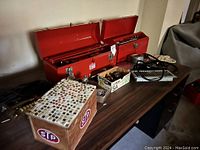 Photo showing two red metal toolboxes open and displaying an assortment of hand tools, tape measures, gloves, an STP box, and a cassette tape on a wooden surface.