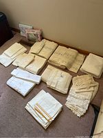 Overview of all linens arranged on floor, showing pile of folded cream and white sheets and pillow shams, some with lace and embroidery details.