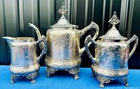 Full set of ornate silverplated coffee pot, milk jug, and sugar bowl on blue surface with black panel background