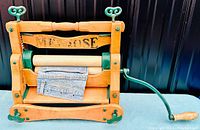 Side view of the wooden and green iron clothes wringer with hand crank and wooden rollers, showing wear and the brand marking.