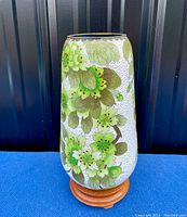 Frontal view of Asian cloisonné vase showing white enamel with green leaves and yellow flowers with brass rim and wooden stand beneath.